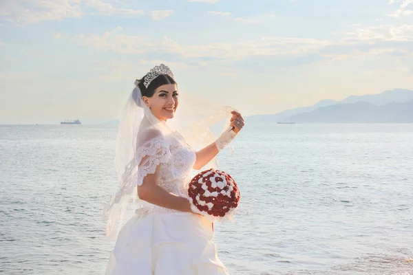 Wedding by the sea. Bride with a red bouquet. Mountains and sea background. Adorable bride. Sunrise view. Concept marriage, just married.