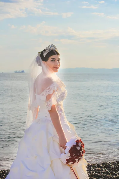 Wedding by the sea. Smilling bride with a red bouquet. Mountains and sea background. Adorable caucasian bride. Concept marriage, just married