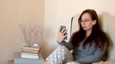 Bir kadın evde telefonla video görüşmesi yapıyor. Gözlüklü ve kulaklıklı kız yatakta akıllı telefonla konuşuyor..