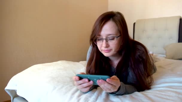 リラックスした若い女性の笑顔は リビングルームでベッドの上でビデオを見てスマートフォンを保持します 幸せな女の子とともに眼鏡見ます携帯電話 — ストック動画