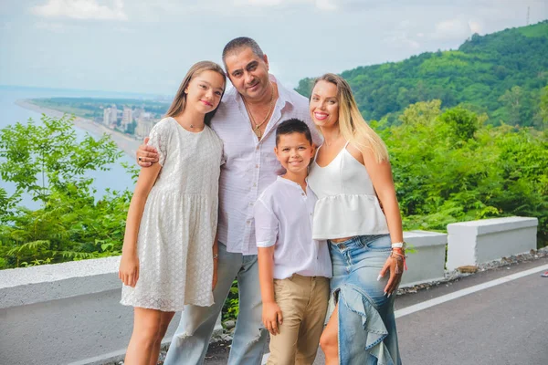 Neşeli, mutlu aile, baba, anne, oğul ve kız deniz kenarında yürüyüş yapmaktan zevk alıyorlar. Yürüyüş yolunda beyaz elbiseli bir aile. Yeşil orman arka planı.