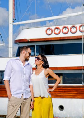 Limanda yürüyen mutlu aşıklar çifti. Arka planda gemi var. Beyaz bluzlu, neşeli, gülümseyen adam ve beyaz bluzlu şanslı bayan ve sarı pantolon, açık deniz limanında..