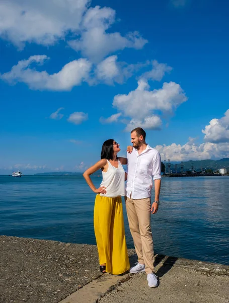 Happy couple of lovers walking at the harbour. Mountains at background. Cheerful, smiling man in white and lady in white blouse and yellow trousers, outdoors at sea port.