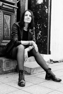 Siyah deri ceketli esmer kadın taş basamakta oturuyor. Rock konsepti. Georgia, Batumi 'deki kız model. Arka plandaki eski kapı. Siyah ve beyaz.