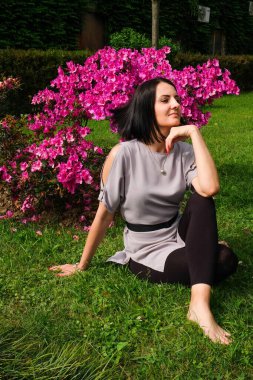 Güzel esmer kadın parkta pembe çiçeklerin yanında çimlerin üzerinde oturuyor. Gülümse, Batumi, Georgia 'daki tatlı bayan. Yaz zamanı..