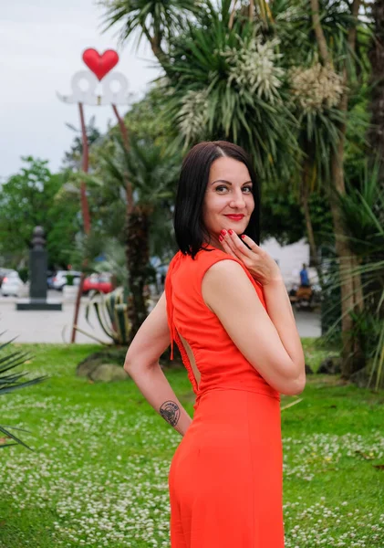 Moda, esmer model, kırmızı takım elbiseli kadın. Seksi, modern tulumlar. Parkta poz veren bir kız, arka planda avuçlar..