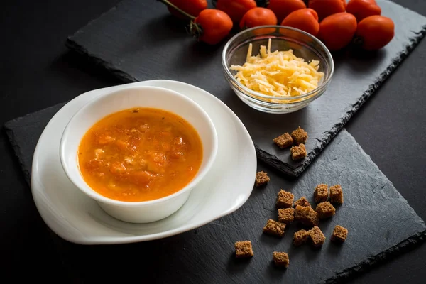 Сливочный томатный суп с сыром на черном деревянном фоне. Тосты за сухой хлеб, помидоры черри, специи и чеснок на заднем плане . — стоковое фото