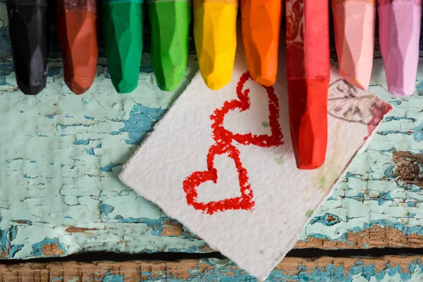 Bright colored pencils on an old blue green wooden background. Two red hearts painted on a slice of paper