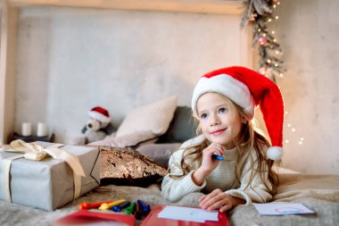 Kırmızı Noel şapkalı küçük kız Noel Baba 'ya mektup yazıyor. Altın Noel ışıkları evin parlak oturma odasında sallanıyor. Noel Hediyesi Bekleniyor