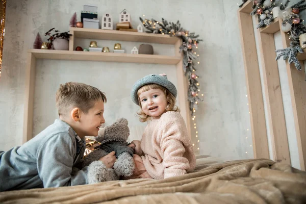 Bratr a sestra v jasném obývacím pokoji doma na posteli. malé děti přátelé v kloboucích, zabalené v teplé přikrývce, hraní s medvídkem na pozadí vánoční výzdoba. — Stock fotografie