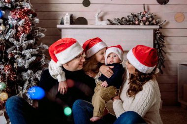 Evde güzel akrabalar, anne ve babasıyla güzel bir kız, kırmızı Noel Baba şapkalı, şöminenin yanındaki ışıklarla süslenmiş Noel ağacı. Kutla, tatil.