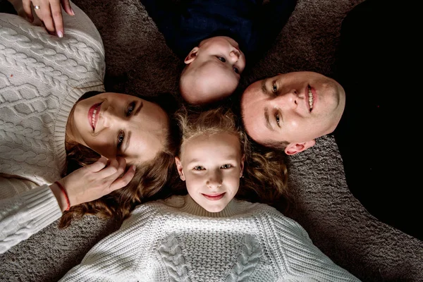 Pohled na krásné mladé rodiče, roztomilá dceruška a syn se drží za ruce, dívají se na kameru a usmívají se, leží na dřevěné podlaze — Stock fotografie