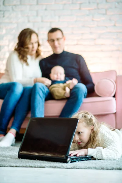 Linda niña está usando una tableta digital mientras está acostada en el suelo en casa, en el fondo, su hermoso padre y madre con el bebé en el sofá. El concepto de bienestar familiar, educación para el cuidado — Foto de Stock