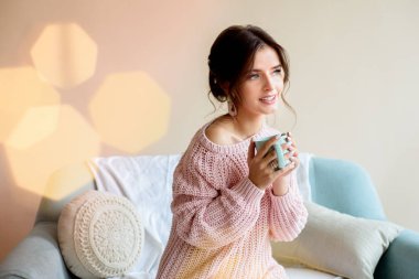 Işık odasındaki rahat bir kanepede bir fincan kahveyle güzel bir kız. Pembe bir kazak ve beyaz çorapla. Noel ışıkları. Samimi mutlu noeller. Bir sürü renkli Bokeh ışıkları