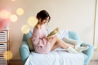 Işık odasındaki rahat bir kanepede bir fincan kahveyle güzel bir kız. Pembe bir kazak ve beyaz çorapla. Noel ışıkları. Samimi mutlu noeller. Bir sürü renkli Bokeh ışıkları