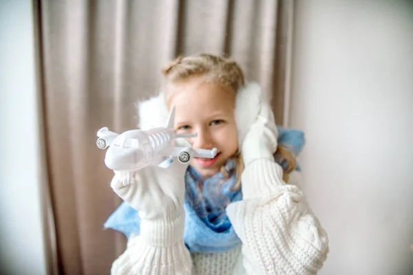A child plays with an airplane, a cozy winter in a knitted sweater and a white fluffy hat. music in fur headphones. Winter travel concept. Winter holiday. Classic blue scarf — Stock Photo, Image