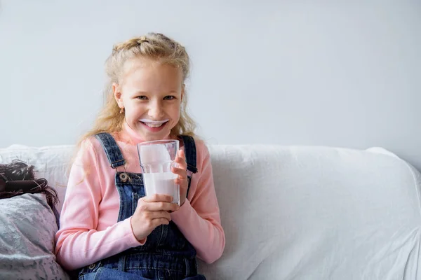 A rapariga bebe kefir ou leite ao pequeno-almoço. bigode engraçado. rindo no sofá em casa. alimentação saudável crianças — Fotografia de Stock