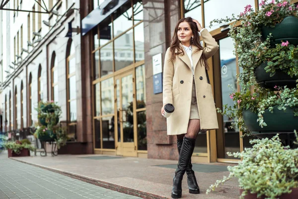 Jeune femme brune en plein air. Photo de rue. Tasse en carton de café dans les mains. Mode d'automne. Manteau beige, jupe à la mode et bottes hautes. Joyeux sourire fille court dans les escaliers . — Photo