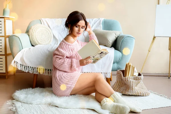 Портрет молодої красивої жінки, що читає книгу на дивані, затишна світла кімната, різдвяний настрій. Темне волосся, в'язаний рожевий светр. Святкові різдвяні гірлянди вогні. Ефект магічного снігопаду . — стокове фото