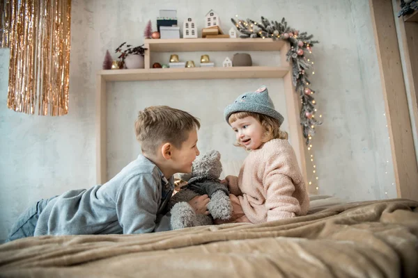 Irmão e irmã na brilhante sala de estar em casa na cama. crianças pequenas amigos em chapéus, envolto em um cobertor quente, brincando com um urso de pelúcia no fundo da decoração de Natal . — Fotografia de Stock