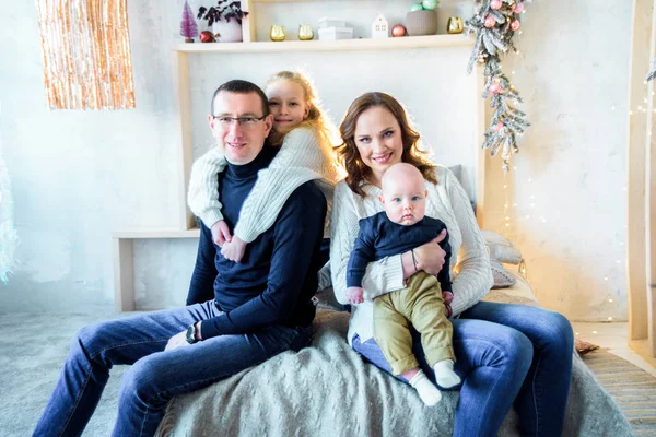 Happy mom and dad, sister and little brother on the bed in a large bright bedroom. Christmas decor and lights. Big happy family. The concept of parental love and parenting