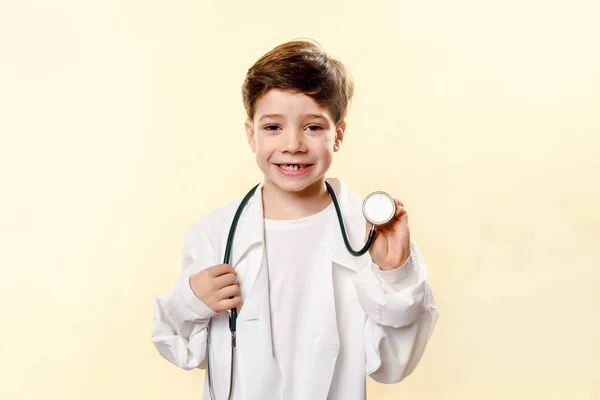 Adorabile futuro dottore su sfondo bianco. Un ragazzo interpreta un dottore. giochi per bambini. Piccolo dottore. cappotto bianco e fonendoscopio. Bambino emotivo In uno studio — Foto Stock
