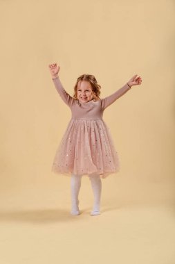 Sarışın, mutlu küçük kız, duygusal olarak şaşırmış. Stülden yapılmış zarif bir elbiseyle stüdyoda pembe bir arka planda. Kıvırcık saçlı. Boşluğu kopyala