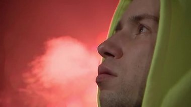 Genç bir adam kırmızı bardaktan sıcak çay ya da kahve içip sigara içiyor. Yüze yakın çekim, sokak ışıkları, sis..