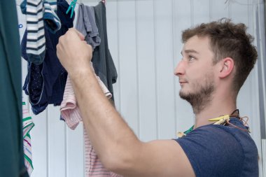 30 yaşlarında, sakallı bir adamın portresi, kıyafetlerini çamaşır ipine asıyor, bekar ve yalnız bir hayat sürüyor, giysi askıları iç çamaşırı bağlıyor, bir kadına yardım ediyor, erkek işi değil..