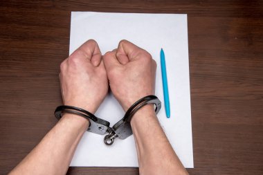 A man with bare hands in handcuffs sits at a table in front of a blank sheet of paper and a fountain pen. Concept: the detainee writes a confession, the prisoner signs the document. clipart