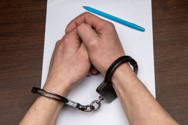 A man with bare hands in handcuffs sits at a table in front of a blank sheet of paper and a fountain pen. Concept: the detainee writes a confession, the prisoner signs the document. clipart