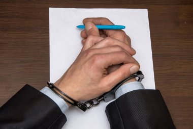 Elleri kelepçeli bir adam boş bir kağıt ve dolma kalemle masada oturuyor. Konsept: Tutuklu itiraf yazar, tutuklu belgeyi imzalar..