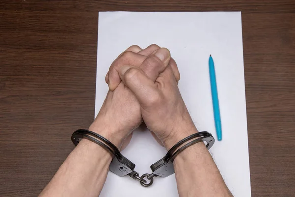 手錠をかけられた男が紙と噴水のペンの前のテーブルに座っている 被収容者は自白を書き 囚人は文書に署名する — ストック写真