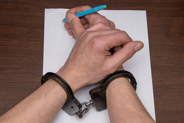 手錠をかけられた男が紙と噴水のペンの前のテーブルに座っている 被収容者は自白を書き 囚人は文書に署名する — ストック写真