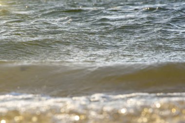 Deniz dalgası kumlu plajın üzerinden geçiyor, güneş ışınlarında parıldıyor. Sonsuz su alanı.