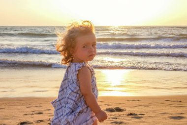 Kumlu bir sahilde batan güneş ve deniz dalgalarının arka planında küçük bir kız kameraya bakar. Ortalama plan, güneş ışığı ve renk aydınlatması..
