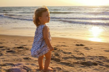 Kumlu bir sahilde küçük bir kız gün batımını seyrediyor, güneşin parıltısını..