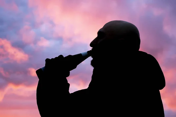 Umriss eines Mannes, der bei Sonnenuntergang Bier aus einer Flasche trinkt, in schwarzen Handschuhen, rosa Wolken bei Sonnenuntergang. — Stockbild Umriss Eines Mannes Der Bei Sonnenuntergang Bier Aus Einer Flasche — Stockfoto