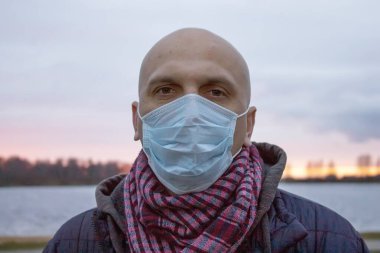 35-40 yaşlarında, gölün arka planında tıbbi maske takmış kel bir adamın sokak portresi. Kavram: Koronavirüsün yayılmasının önlenmesi, önlemler ve kendini izole etme.