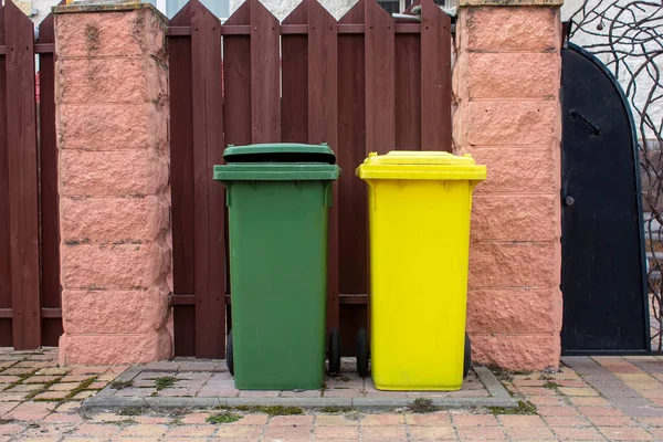 Two plastic garbage containers are in the yard near the fence, garbage collection.