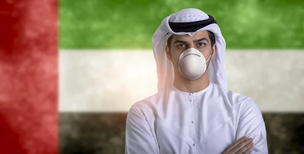 Emirati 'li genç adam kendini koronavirüsten korumak için maske takıyor BAE bayrağının önünde duruyor.