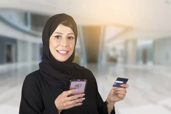 Abaya 'daki zarif Arap kadın elinde cep telefonu ve plastik kartla alışveriş merkezinde dikiliyor. Şık tesettürlü iş kadını cep telefonuyla alışveriş yaparken kredi kartıyla ödeme yapıyor..