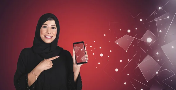 Arap mutlu, tesettürlü, boş ekran cep telefonu tutan ve kırmızı arka planı işaret eden genç bir kadın. Arap teknolojisi kavramı.