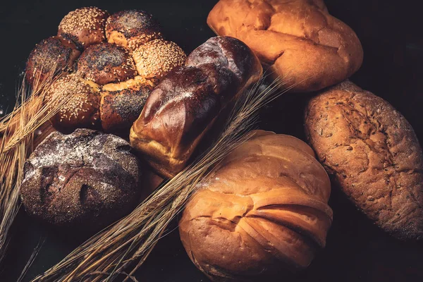 Pekařský chleba na dřevěném stole. Různý chléb a svazek pšeničných uší. Fotografie v tmavém klíči. — Stock fotografie