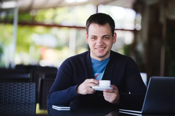 Красивый молодой человек работает на ноутбуке и улыбается, наслаждаясь кофе в кафе — стоковое фото