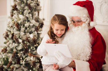 Küçük kız koltukta evde tablet kullanarak Santa ile oturma odası oturma