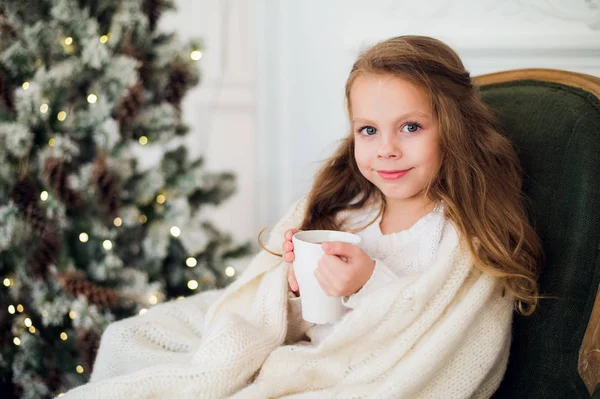 Het dragen van pyjama zittend op fauteuil gewikkeld in een deken met vakantie beddengoed door kerstboom en open haard consumptiemelk thuis meisje — Stockfoto