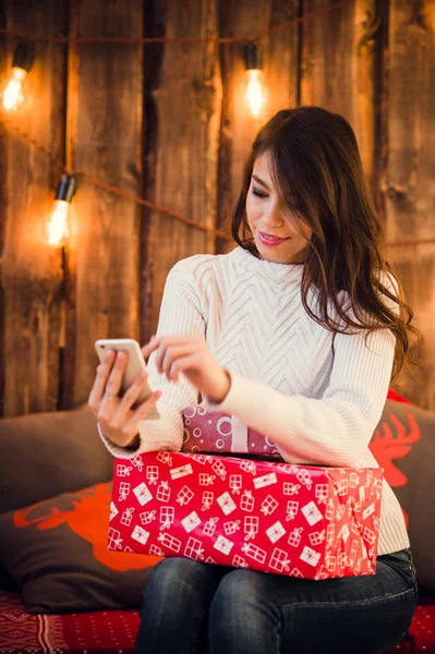 Νεαρή γυναίκα που κάθεται στο πάτωμα χρησιμοποιώντας κινητό τηλέφωνο μηνύματα κοντά διακοσμημένα Χριστούγεννα τοίχο με λαμπτήρες. — Φωτογραφία Αρχείου