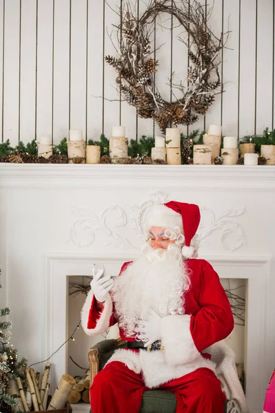 Felice Babbo Natale digitando un messaggio sul cellulare e sorridendo seduto sulla sua sedia con camino. Albero di Natale sullo sfondo — Foto Stock