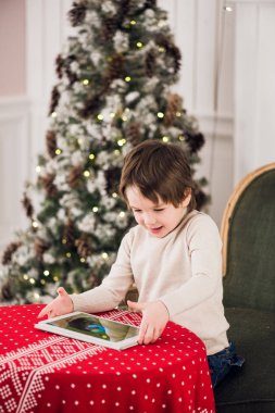 Yeşil sandalyede oturan ve Noel zamanı sırasında bilgisayar tablet ile oynayan sevimli çocuk portresi. Tatil mevsim.